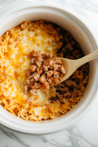 This image shows the cooked sausage being layered over the hash browns in the crockpot, adding a savory depth to the casserole.