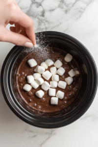 This image shows mini marshmallows being sprinkled on top of the freshly made hot chocolate in the crockpot, adding a soft, sweet touch to the warm beverage.