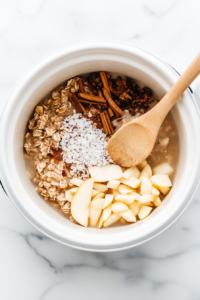 This image shows oats, diced apples, and warm spices being added to the crockpot, setting the base for a comforting and hearty oatmeal.