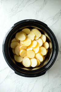 This image shows the first layer of thinly sliced potatoes being evenly arranged at the bottom of the Crockpot, ready to be coated with the creamy sauce.