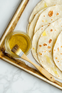 This image shows tortillas being neatly placed on a baking sheet, ready to be brushed with oil and filled with ingredients.