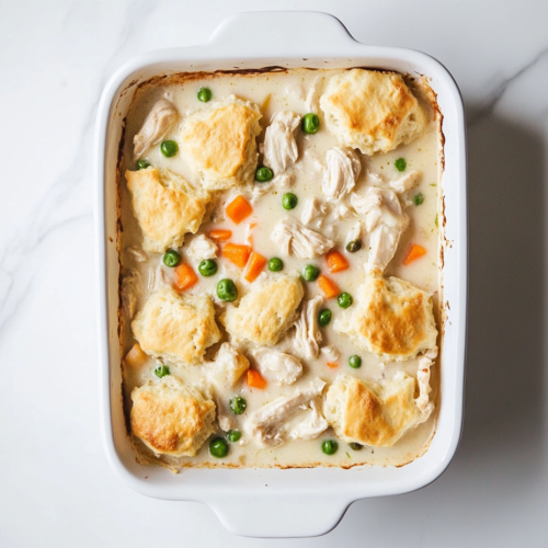 This image shows a freshly baked Chicken Biscuit Bake in a white rectangular dish, filled with a creamy chicken mixture, peas, and carrots, topped with golden biscuits.