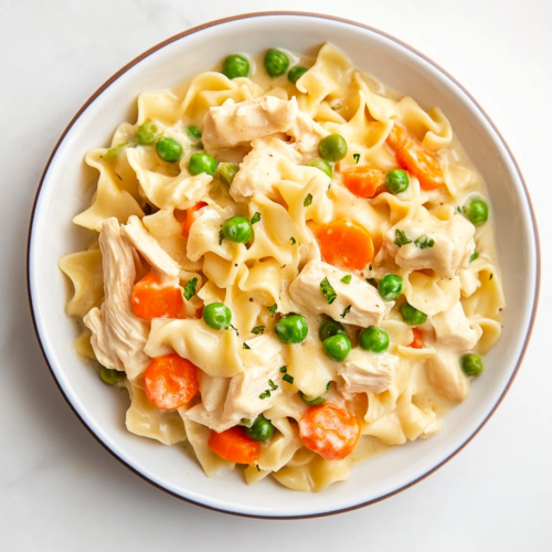 This image shows a Chicken Pot Pie Noodle Skillet served on a round white plate, featuring tender noodles, shredded chicken, bright carrots, and peas coated in a creamy sauce.