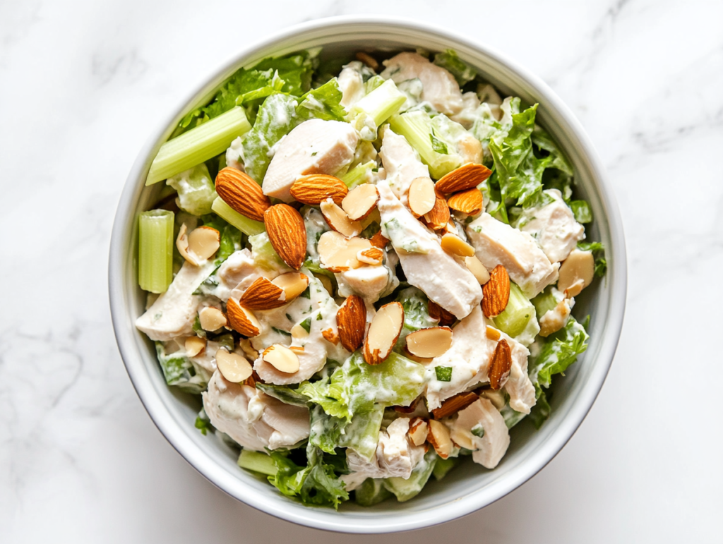 This image shows a white bowl filled with creamy Chicken Salad, made with cooked shredded or chopped chicken, toasted almonds, and chopped celery, creating a fresh and crunchy texture.