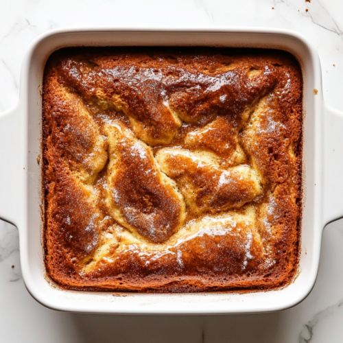 This image shows a freshly baked Cinna-Bun Cake in a square baking dish, perfectly golden brown with a visible cinnamon swirl, ready to be served.