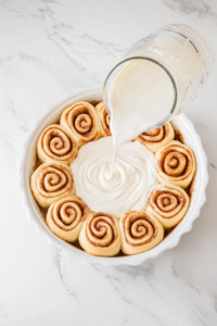 This image shows cinnamon rolls neatly arranged in a baking dish as rich heavy cream is poured over them, ensuring a soft and gooey texture.