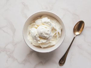 This image shows a scoop of smooth, creamy vanilla ice cream in a white bowl, with a golden-colored spoon resting beside it, ready to be enjoyed.