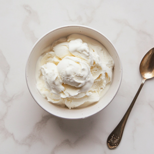 This image shows a scoop of smooth, creamy vanilla ice cream in a white bowl, with a golden-colored spoon resting beside it, ready to be enjoyed.