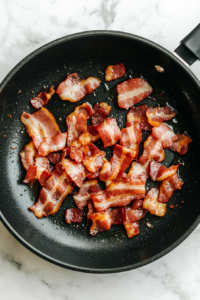 This image shows small pieces of bacon being cooked in a pan until crispy, releasing their smoky and savory aroma to enhance the dish.