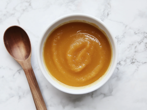 This image shows a small round white bowl filled with homemade Crockpot applesauce made with apples and pumpkin pie spice, with a spoon placed beside it for serving.