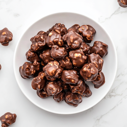 This image shows a white plate piled high with rich, chocolatey crockpot peanut clusters, with additional clusters scattered around the plate, ready to be enjoyed as a decadent dessert.