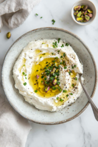 This image shows a well being created in the center of the whipped feta, filled with golden honey and garnished with fresh chives, pistachios, and black pepper.