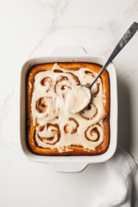 This image shows a spoon drizzling a creamy vanilla glaze over the warm Cinna-Bun Cake, adding a glossy, sweet finishing touch.