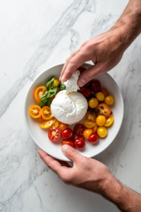 This image shows hands gently tearing apart a ball of burrata cheese, ensuring a creamy, rich texture that will blend beautifully with the juicy tomatoes in the salad.