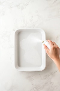 This image shows a rectangular baking dish being greased with cooking spray to prevent sticking and ensure easy serving.