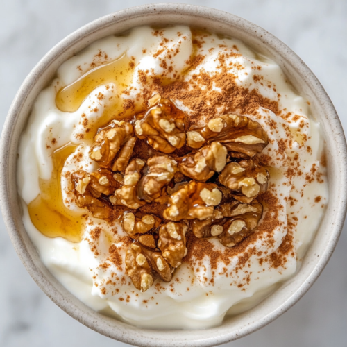 This image shows a bowl of creamy Greek yogurt topped with golden honey drizzle and chopped walnuts, creating a simple yet delicious dessert with a rich and nutty flavor.