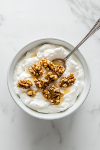 This image shows a dusting of cinnamon powder being sprinkled over the yogurt and honey-coated walnuts, adding a warm, aromatic touch to the dessert.