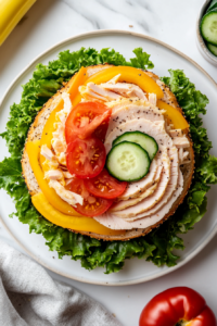 This image shows the process of assembling the turkey bagel sandwich by layering turkey, cheese, vegetables, and mustard on the bottom half of the bagel before topping it with the other half.