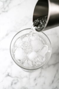 This image shows gin, vodka, and Lillet Blanc being poured into a mixing glass over ice, blending the key ingredients for the Vesper cocktail.