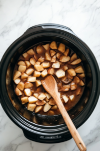 This image shows a hand stirring chopped apples, spices, and liquid in the slow cooker to ensure even distribution of flavors before cooking.