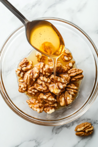 This image shows warm toasted walnuts being coated in golden honey, creating a glossy and sweet topping for the yogurt dessert.