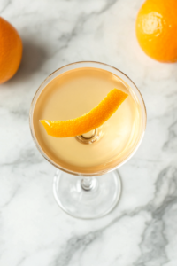 This image shows a delicate orange twist being placed on the rim of the glass, adding a citrusy aroma and an elegant touch to the Vesper cocktail.