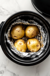 This image shows several foil-wrapped potatoes being arranged inside a slow cooker, ready to be cooked slowly for soft and fluffy baked potatoes.