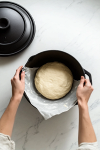 This image shows a round loaf of bread dough being carefully placed into a hot Dutch oven lined with parchment paper, ready for baking.