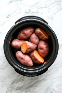 This image shows whole, unpeeled sweet potatoes being placed inside a slow cooker, ready to be slow-cooked until soft and tender.