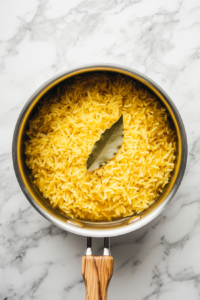 This image shows chicken broth being poured into a pan filled with rice, with a bay leaf floating on top, infusing the dish with extra flavor.