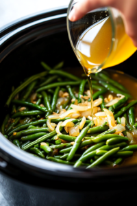 This image shows chicken broth being poured over the green beans in the slow cooker, infusing them with rich, savory goodness.