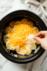 This image shows the creamy mixture being poured over the first layer of potatoes, followed by a generous sprinkle of cheese to add richness to the scalloped potatoes.