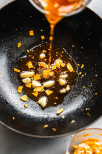 This image shows a vibrant orange sauce mixture being poured into the wok, ready to be simmered and thickened into a delicious glaze.