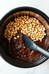 This image shows a generous amount of peanuts being poured into the slow cooker filled with melted, silky chocolate, preparing for a perfect mix.