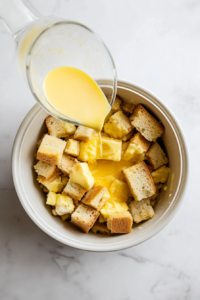 This image shows the creamy egg mixture being poured over the cubed bread in the crockpot, soaking each piece for a flavorful, custardy texture.