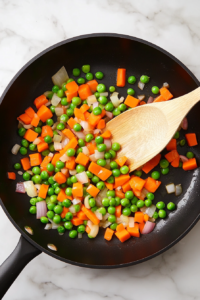 This image shows chopped carrots, peas, and minced garlic being sautéed in a skillet, adding flavor and color to the dish.