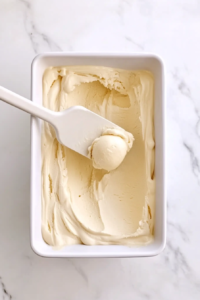 This image shows a spatula transferring freshly churned vanilla ice cream into an airtight container, getting it ready for freezing.