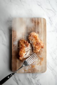 This image shows tender, cooked chicken being shredded with forks on a cutting board, preparing it to be added back into the soup.