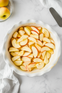 This image shows a pie plate greased with butter, filled with a layer of freshly sliced apples, ready to be transformed into a delicious crustless apple pie.