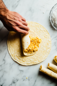 This image shows a tortilla being filled with seasoned mashed potatoes and carefully rolled into a taquito shape, ready for baking.