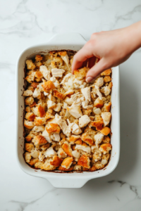 This image shows the buttery stuffing mixture being sprinkled generously over the creamy chicken casserole, ready to create a crispy, golden topping.