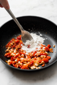 This image shows flour being sprinkled over the sautéed tomatoes and garlic, helping to thicken the sauce for a smooth and creamy shrimp rigatoni.