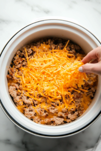 This image shows shredded cheese being generously sprinkled over the sausage layer in the crockpot, ready to melt into gooey perfection.