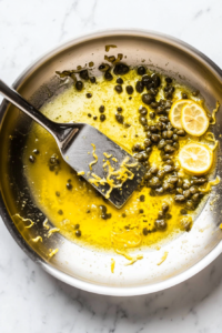 This image shows fresh lemon juice, briny capers, and butter being mixed into the sauce, adding bright and rich flavors.