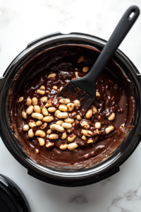 This image shows a hand stirring peanuts into the smooth, melted chocolate inside a slow cooker, ensuring each peanut is fully coated for the perfect peanut cluster.