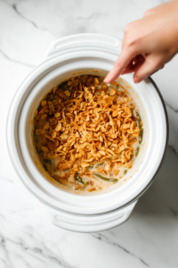 This image shows a generous layer of crispy French fried onions being sprinkled over the top of the cooked green bean casserole, adding a golden, crunchy finish.