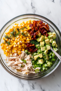 This image shows a bowl filled with cooked corn, chopped green onions, crispy bacon bits, and fresh dill being added to the salad.