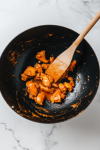 This image shows golden fried chicken pieces being coated in a glossy, sticky orange sauce, ensuring every bite is packed with flavor.