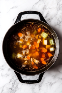 This image shows a pot of broth at a rolling boil, infused with soy sauce, sesame oil, ginger, garlic, carrots, and celery.