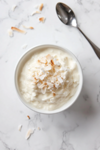 This image shows a bowl of coconut pudding, rich and creamy, topped with shredded coconut for added texture, with a spoon placed beside it, ready for serving.